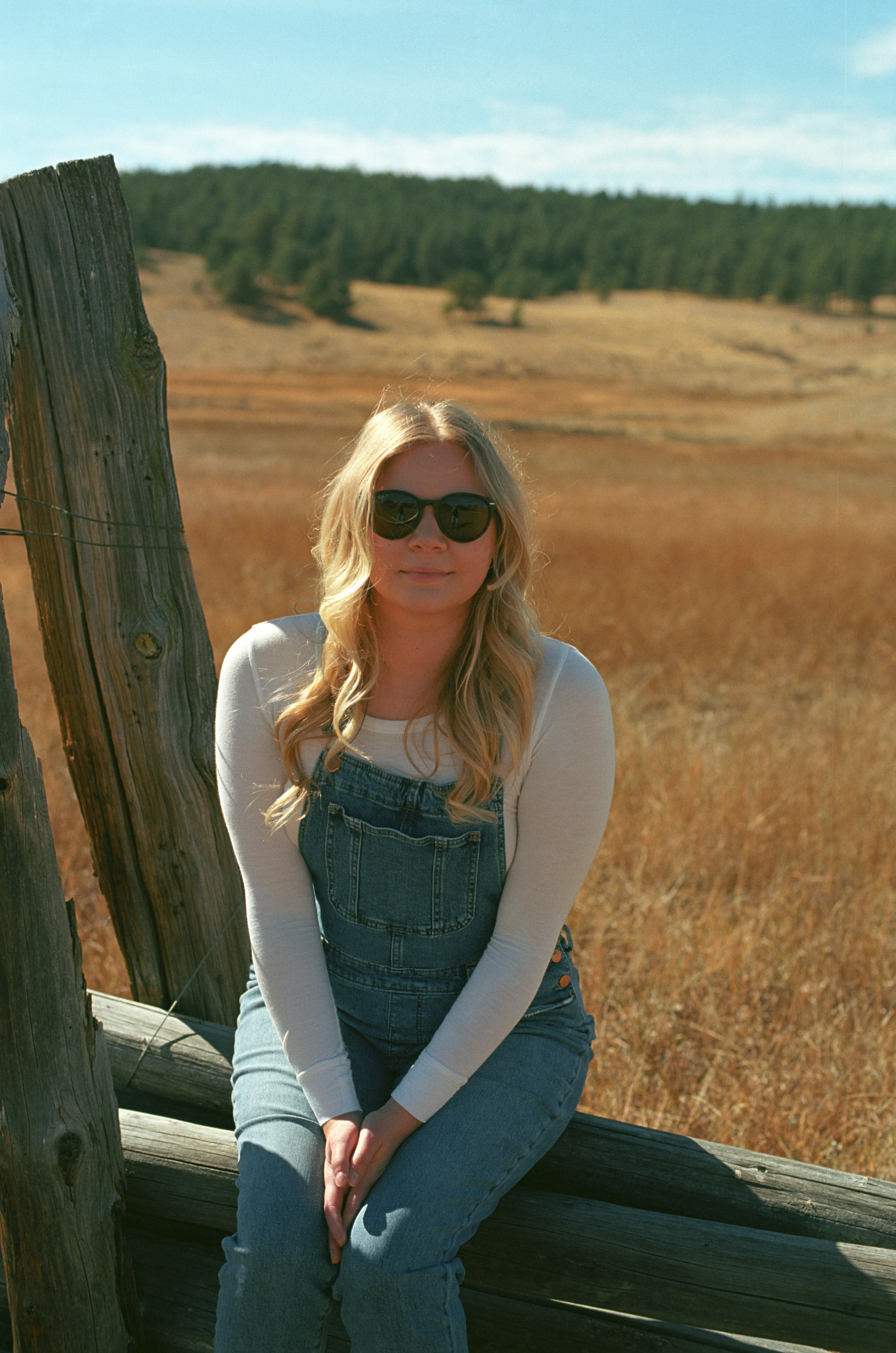 My wife Blake, wearing sunglasses, sitting on a wooden fence in front of a field of grass