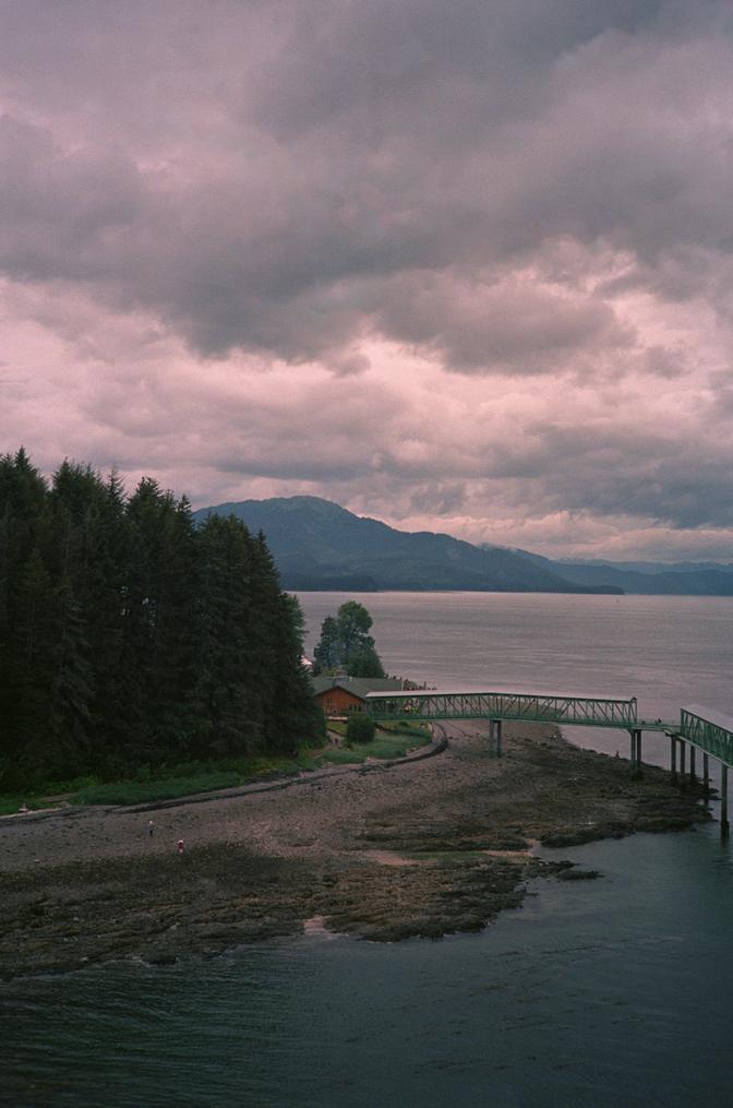 A small brown building on the Alaskan coast nestled between a forest and beach