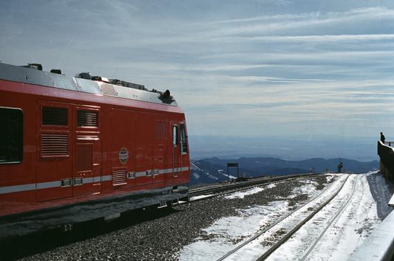 A red train engine parked in the snow at the summit of Pikes Peak, overlooking the city